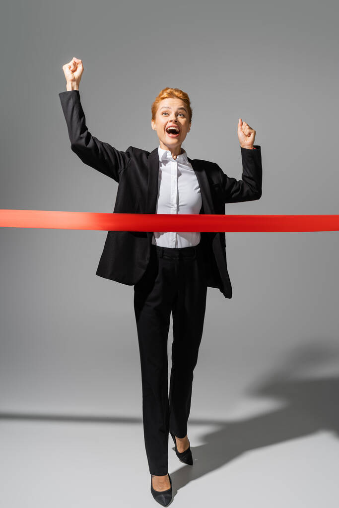 full length of overjoyed businesswoman in black suit crossing finish ribbon on grey background - Photo, Image