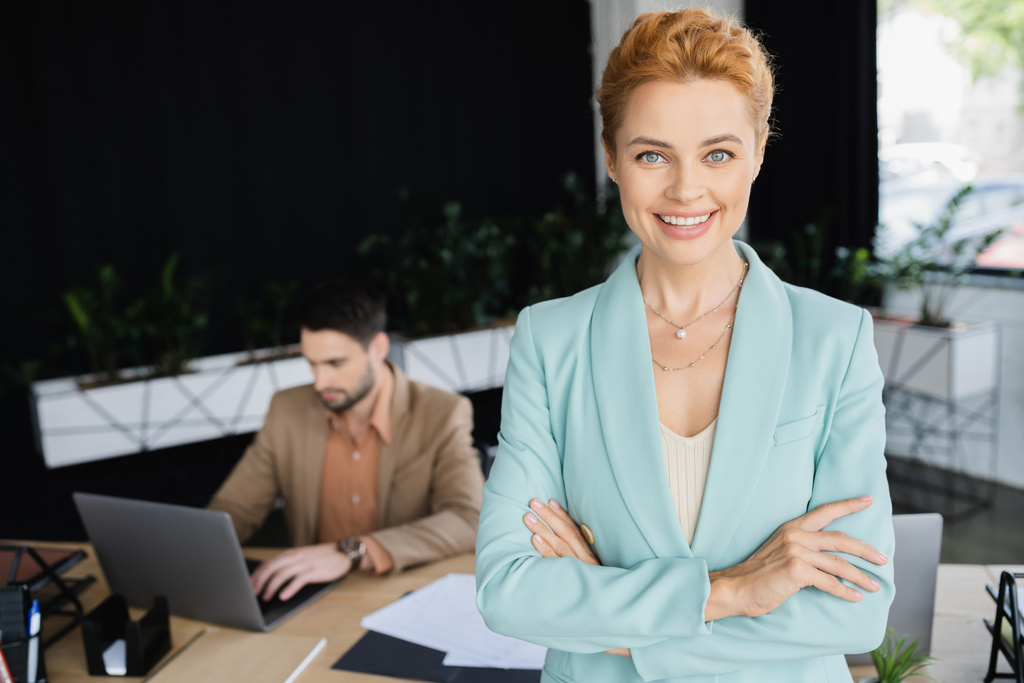 довольная деловая женщина в модном блейзере, смотрящая в камеру рядом с коллегой, работающим на ноутбуке в офисе - Фото, изображение