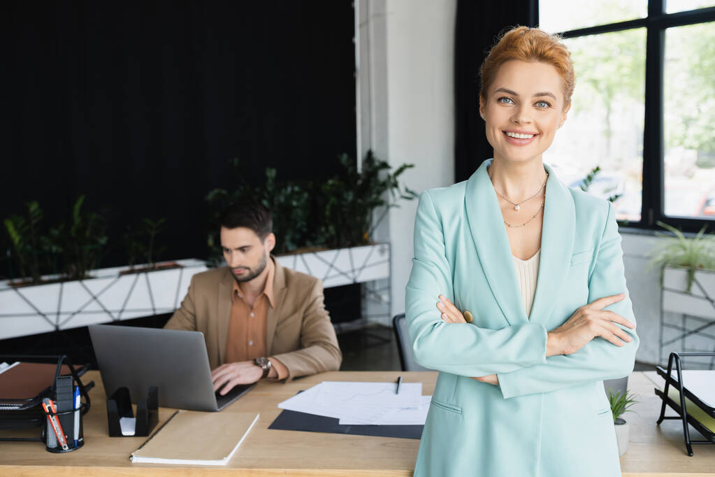 pelirroja mujer de negocios con los brazos cruzados sonriendo a la cámara cerca de colega que trabaja en el ordenador portátil en la oficina - Foto, Imagen