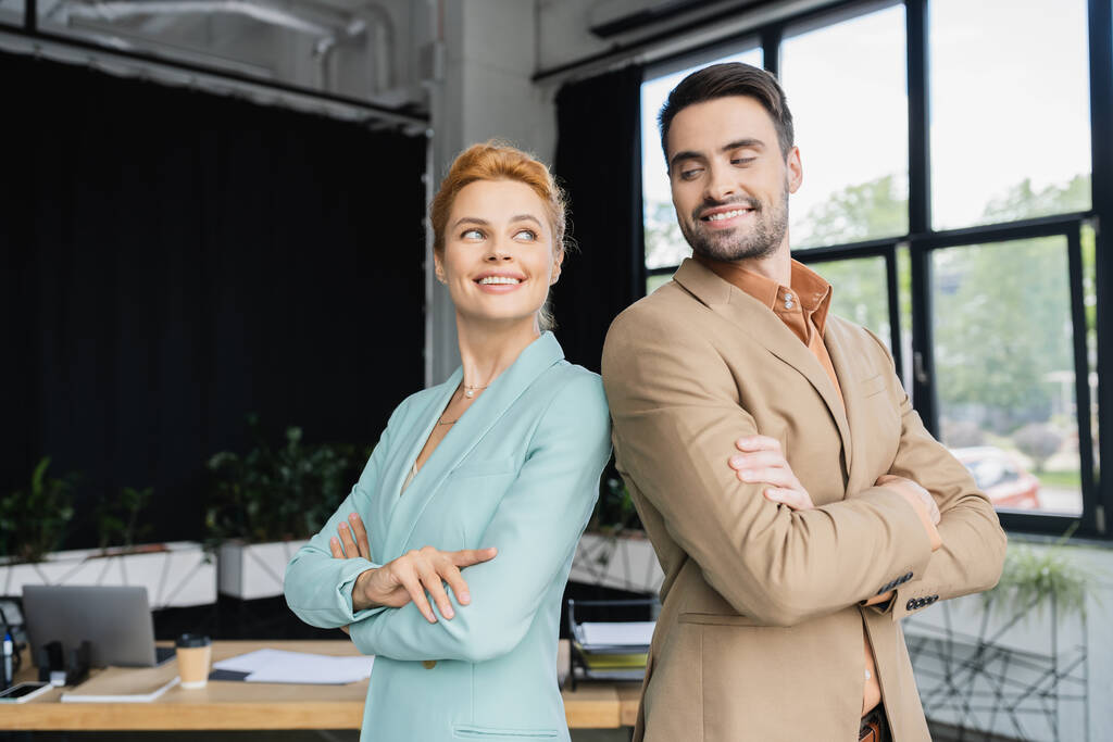 веселые и стильные деловые коллеги, стоящие со сложенными руками и улыбающиеся в офисе - Фото, изображение