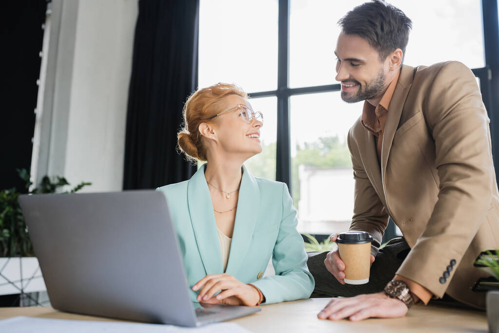 mulher de negócios feliz em óculos trabalhando no laptop e olhando para colega barbudo com café para ir no escritório - Foto, Imagem
