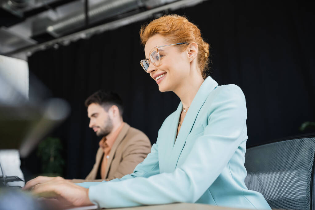 felice donna d'affari rossa in occhiali da vista che lavora vicino a collega su sfondo sfocato in ufficio - Foto, immagini