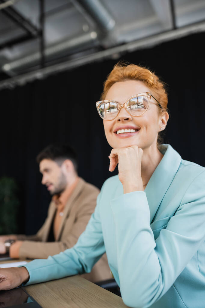 happy and dreamy businesswoman in eyeglasses looking away near blurred colleague working in office - Photo, Image