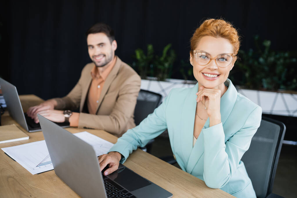 joyful businesswoman smiling at camera near laptop and blurred colleague in office - Photo, Image