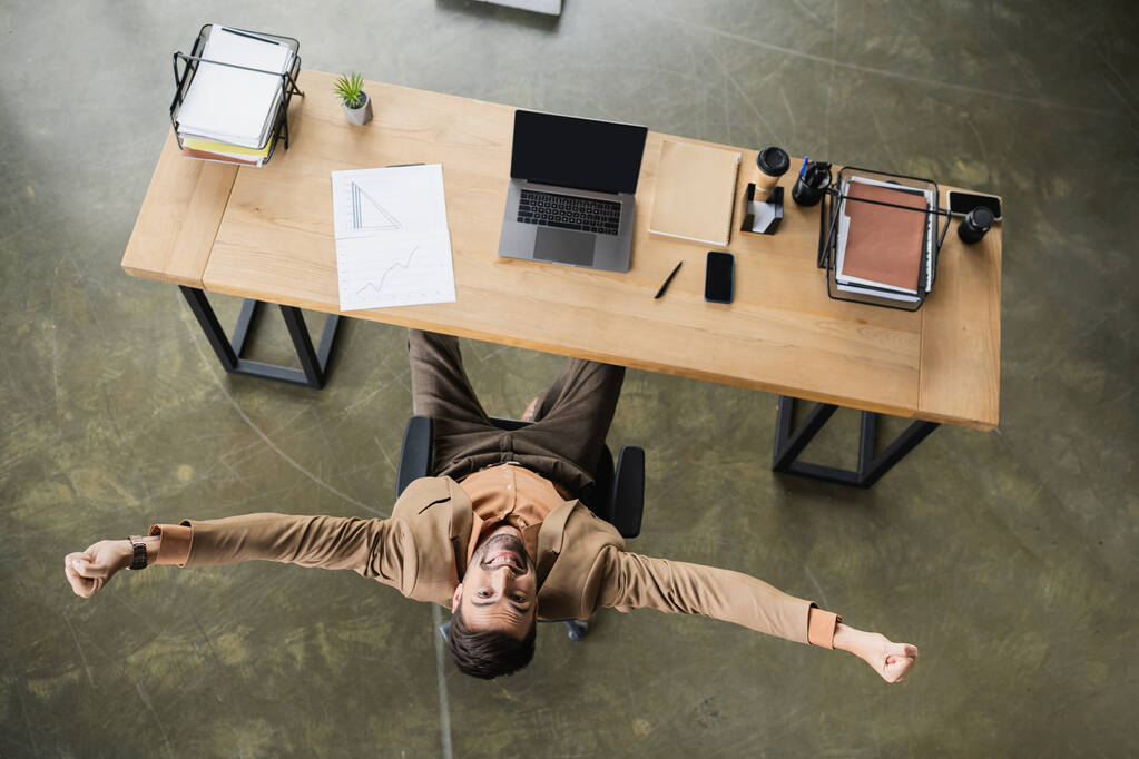 top view of overjoyed businessman showing win gesture near devices and documents on desk in office - Photo, Image