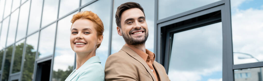 heureux et élégant partenaires d'affaires souriant à la caméra près du bâtiment en verre sur la rue urbaine, bannière - Photo, image