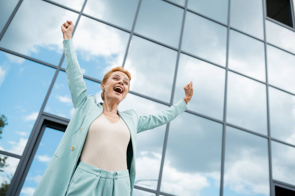 vista de ángulo bajo de la mujer de negocios llena de alegría que muestra el gesto de ganar y gritando cerca del edificio con fachada de vidrio - Foto, imagen