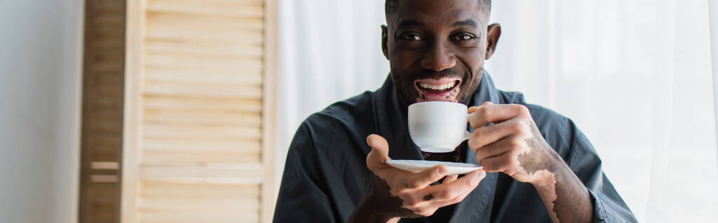 Positivo hombre afroamericano con vitiligo sosteniendo la taza de café y mirando a la cámara en casa, pancarta  - Foto, imagen