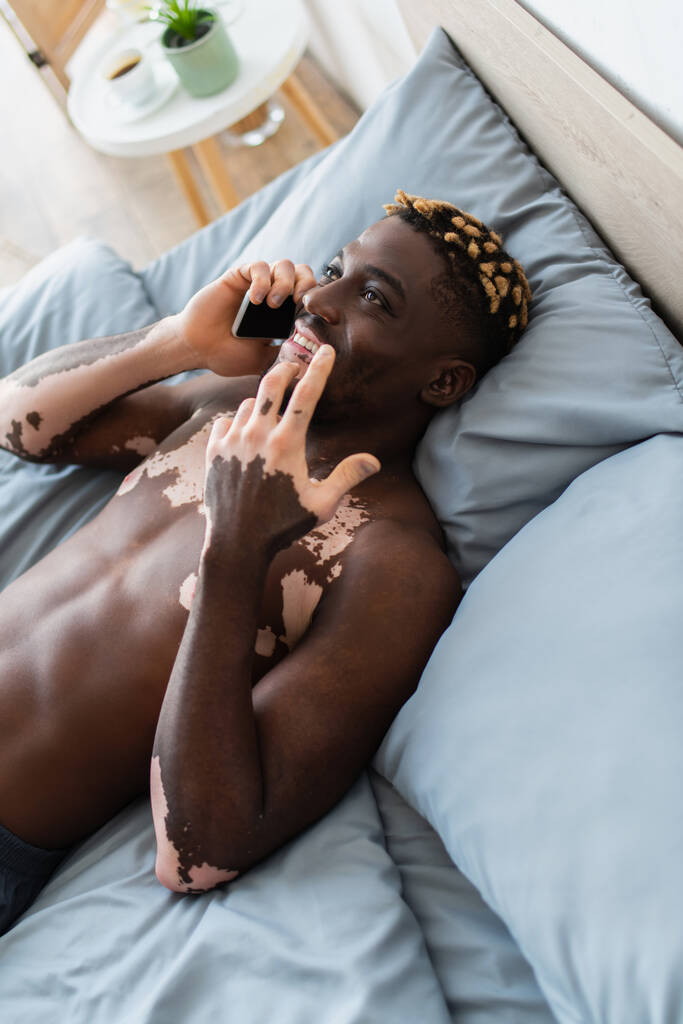 High angle view of cheerful african american man with vitiligo talking on smartphone on bed  - Photo, Image