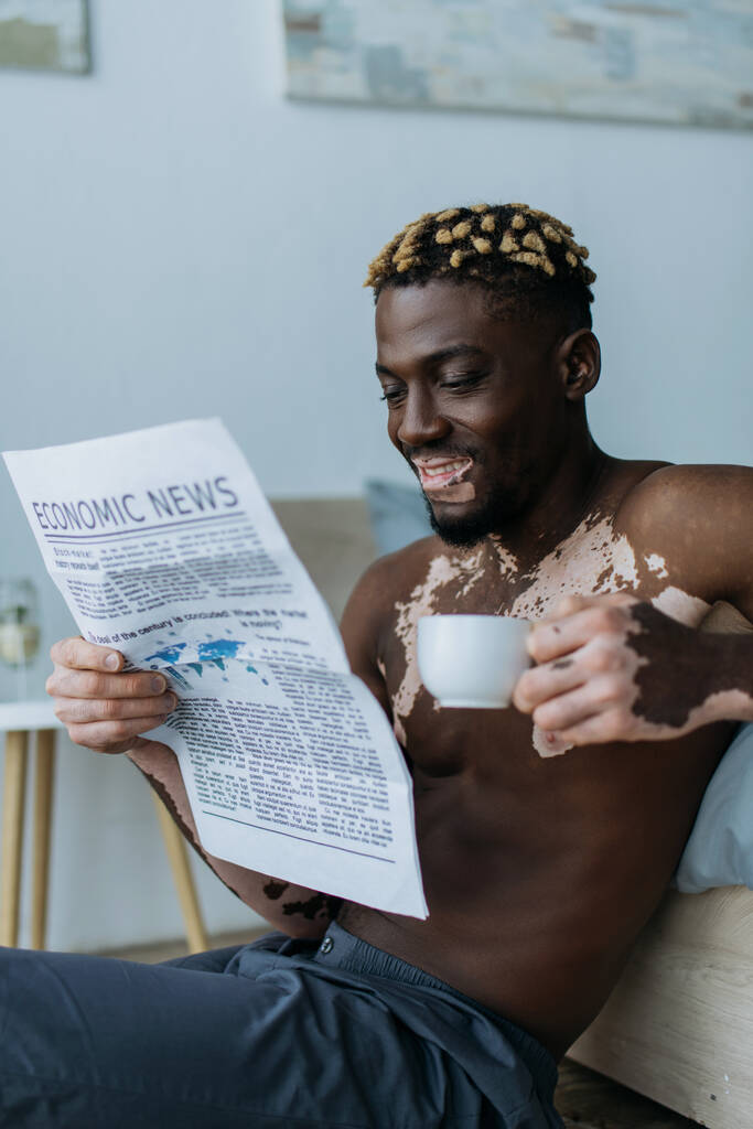 Uśmiechnięty Afroamerykanin z witiligo czytający ekonomiczną gazetę i trzymający kawę w sypialni  - Zdjęcie, obraz