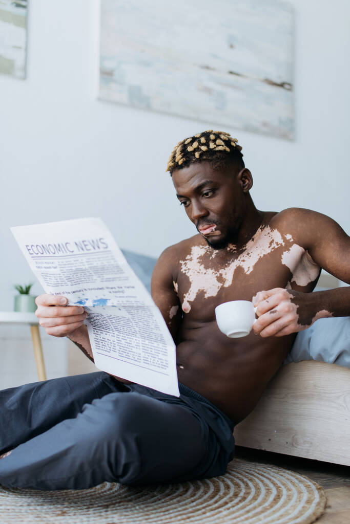 Muskulöser afrikanisch-amerikanischer Mann mit Vitiligo, der im Schlafzimmer Kaffee hält und Zeitung liest  - Foto, Bild
