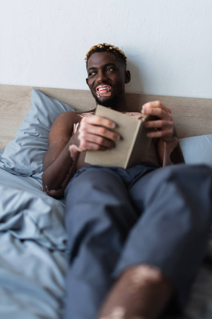 hombre afroamericano sin camisa y sonriente con vitiligo sosteniendo libro mientras está acostado en la cama en casa  - Foto, imagen