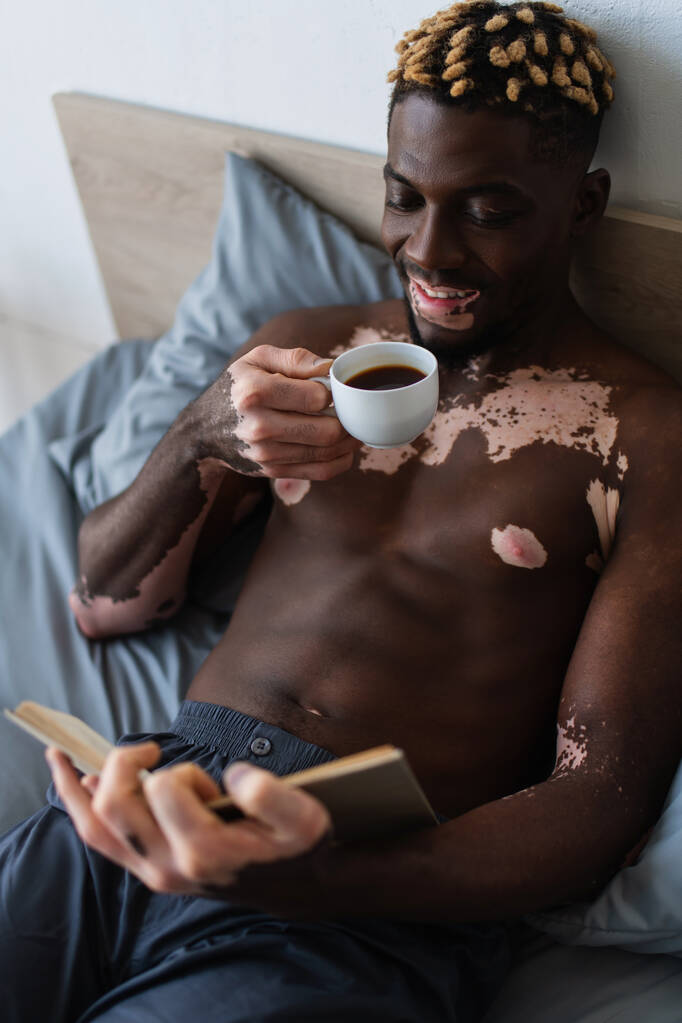 hombre afroamericano sin camisa con vitiligo sonriendo mientras sostiene café y libro de lectura  - Foto, imagen