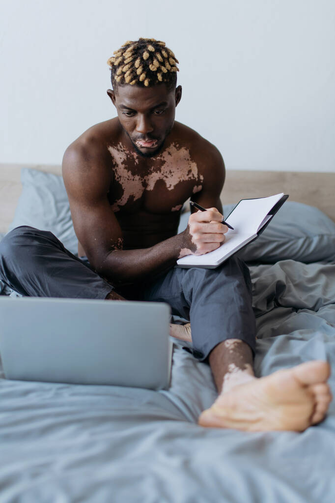Shirtless Afrikaanse Amerikaanse man met vitiligo schrijven op notebook in de buurt laptop op bed thuis  - Foto, afbeelding