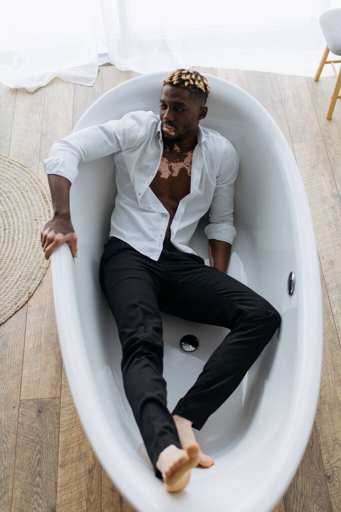 Κάτοψη του Αφροαμερικανού άνδρα με λεύκη σε πουκάμισο και παντελόνι που κάθεται στην μπανιέρα στο σπίτι  - Φωτογραφία, εικόνα