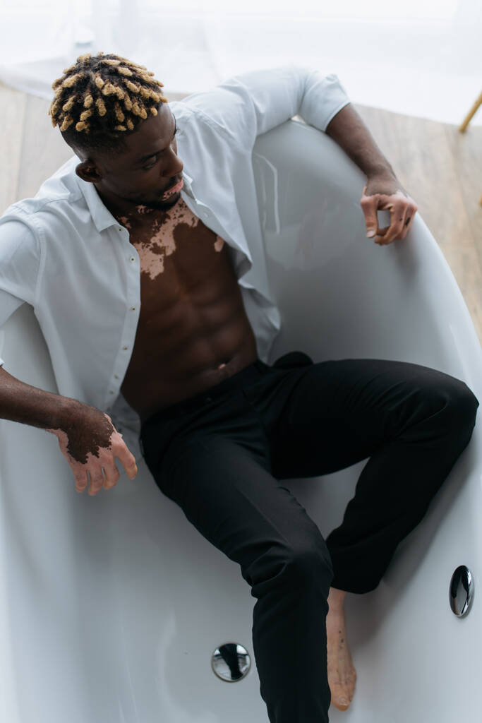 Vista aérea del hombre americano africano muscular con vitiligo sentado en la bañera en casa  - Foto, Imagen