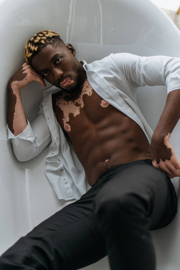 Зверху - мускулястий афроамериканець з вітиліго у сорочці та штанях, що лежали вдома у ванні.  - Фото, зображення
