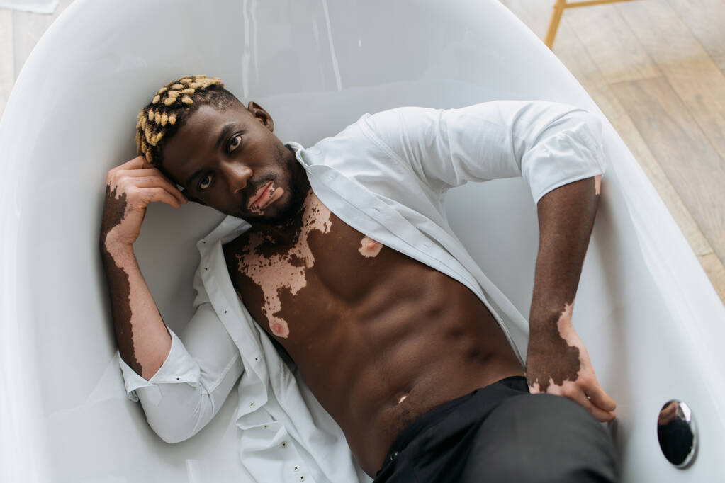 Draufsicht auf muskulöse afrikanisch-amerikanische Mann mit Vitiligo Blick auf Kamera aus der Badewanne zu Hause  - Foto, Bild
