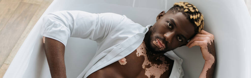 Κάτοψη του Αφροαμερικανού άνδρα με λεύκη σε πουκάμισο που βρίσκεται στην μπανιέρα στο σπίτι, πανό  - Φωτογραφία, εικόνα
