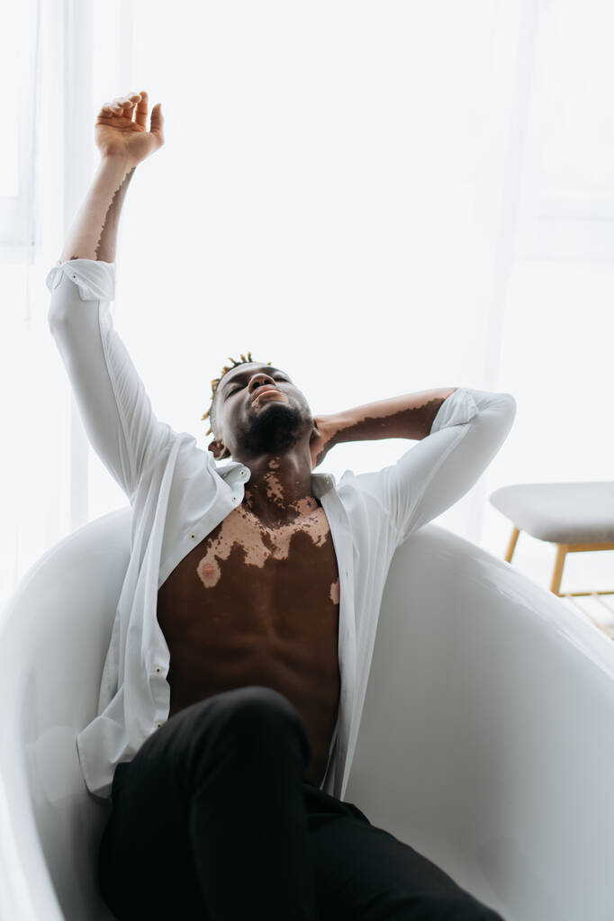 Gespierde Afrikaanse amerikaanse man met vitiligo in shirt zitten in bad thuis  - Foto, afbeelding