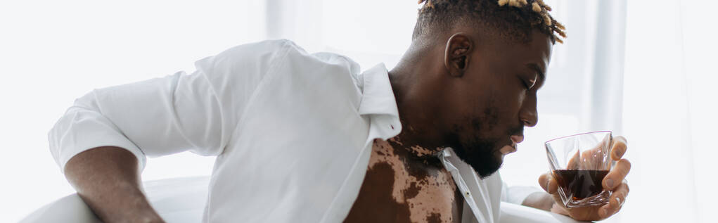 Vista lateral do homem afro-americano com vitiligo em camisa branca segurando uísque enquanto sentado na banheira, banner  - Foto, Imagem