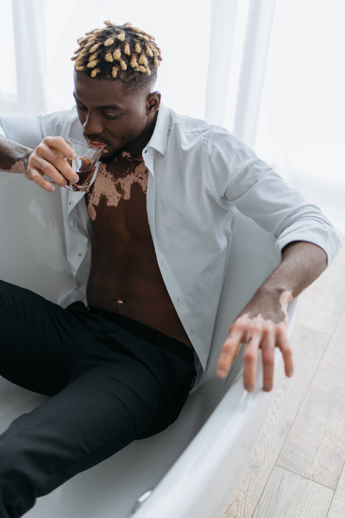 African american man with vitiligo in white shirt holding glass of whiskey while sitting in bathtub  - Photo, Image