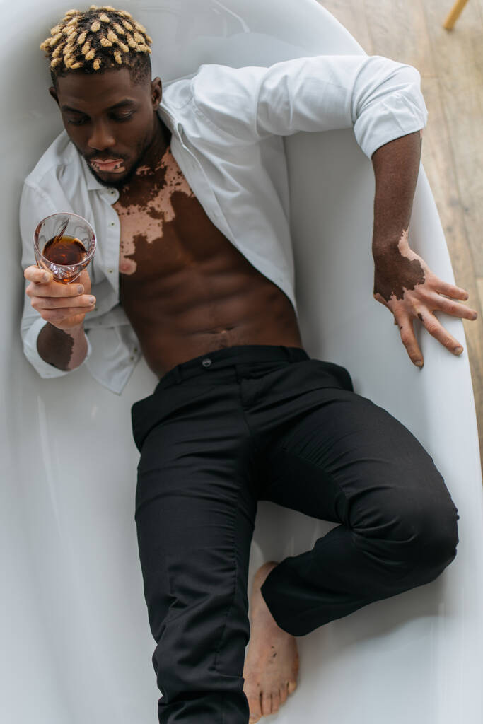 Vista superior do homem afro-americano com vitiligo na camisa e calças segurando uísque na banheira  - Foto, Imagem