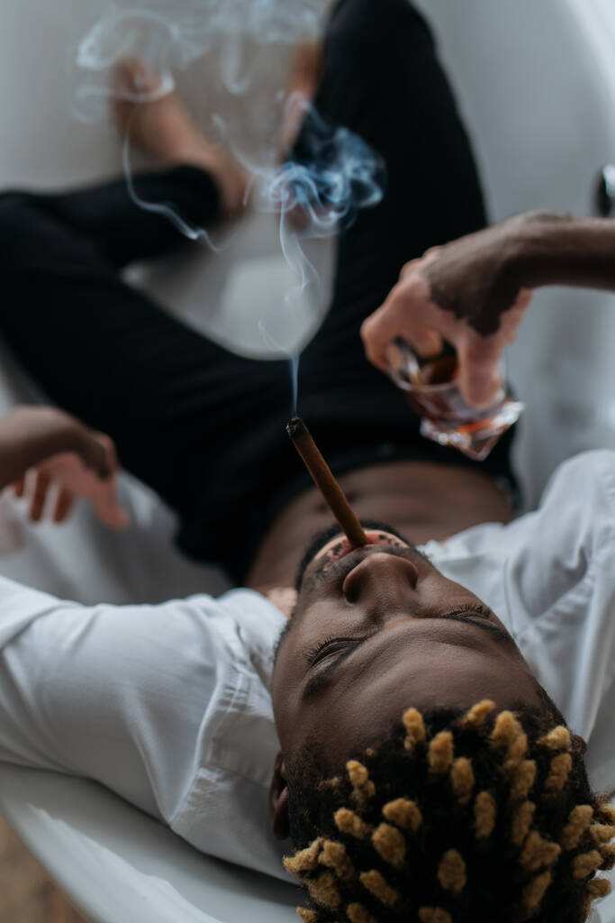 Vista aérea del hombre afroamericano con vitiligo fumando cigarro y sosteniendo whisky en la bañera  - Foto, Imagen
