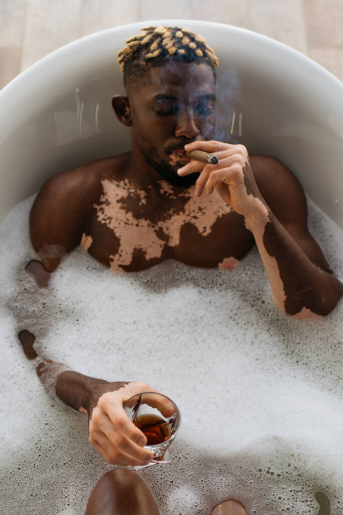 Верхній вигляд африканського американця з вітиліго курить сигару і тримає віскі в ванній з піною.  - Фото, зображення