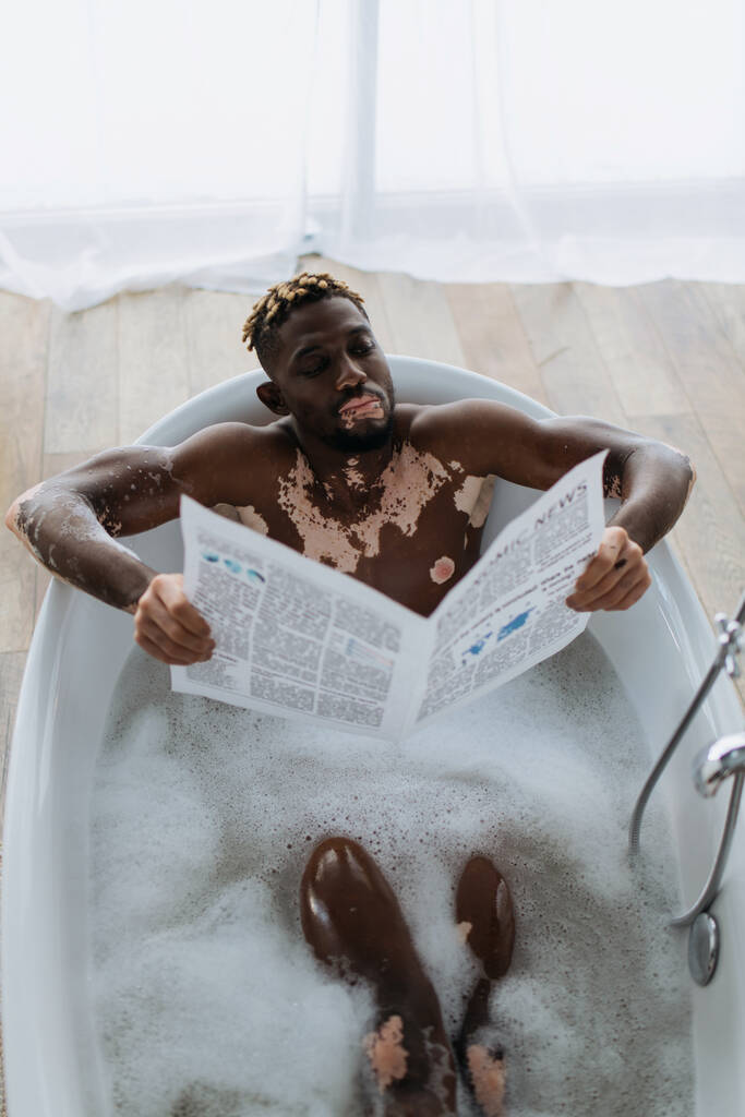 Top view of african american man with vitiligo reading newspaper while taking bath at home  - Photo, Image