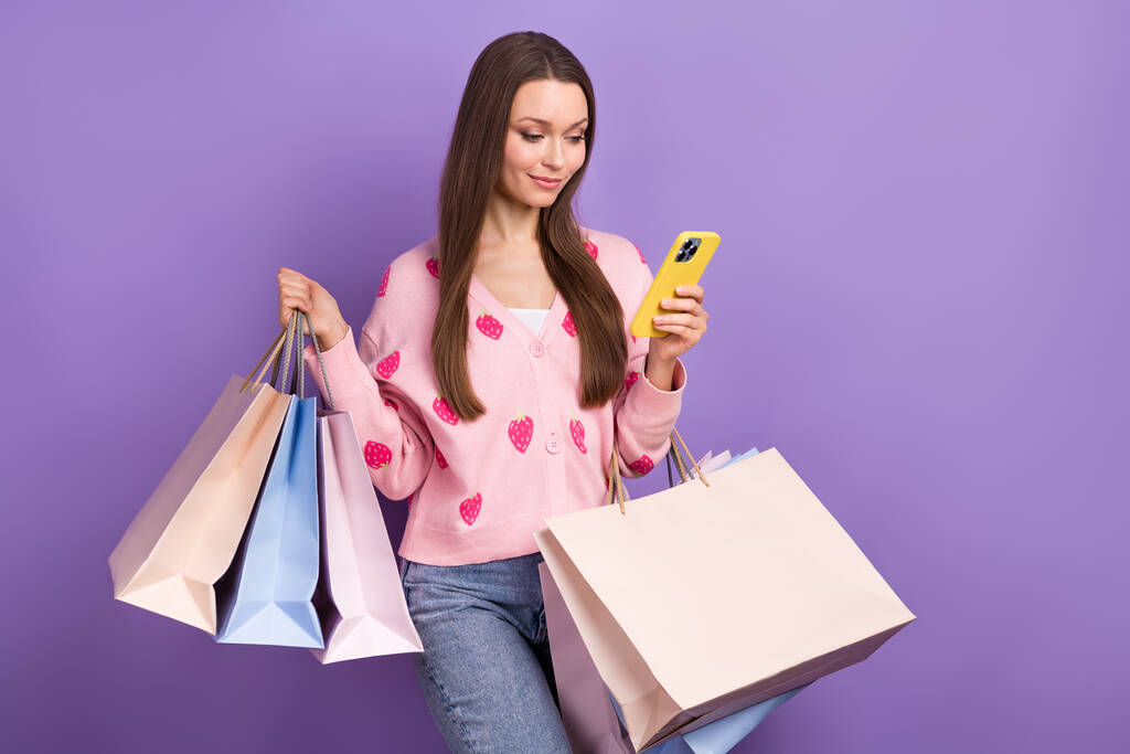 Foto de la joven compradora de moda niña usar vaqueros chaqueta de mezclilla mantener el teléfono navegar oferta en línea bolsas de compras entrega aislada sobre fondo de color púrpura. - Foto, imagen