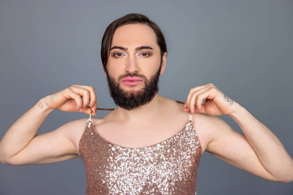 Selbstbewusste ernsthafte transsexuelle Person mit glänzendem Tank-Top, die in die Kamera blickt und seine modische Kleidung berührt, posiert isoliert vor grauem Hintergrund - Foto, Bild