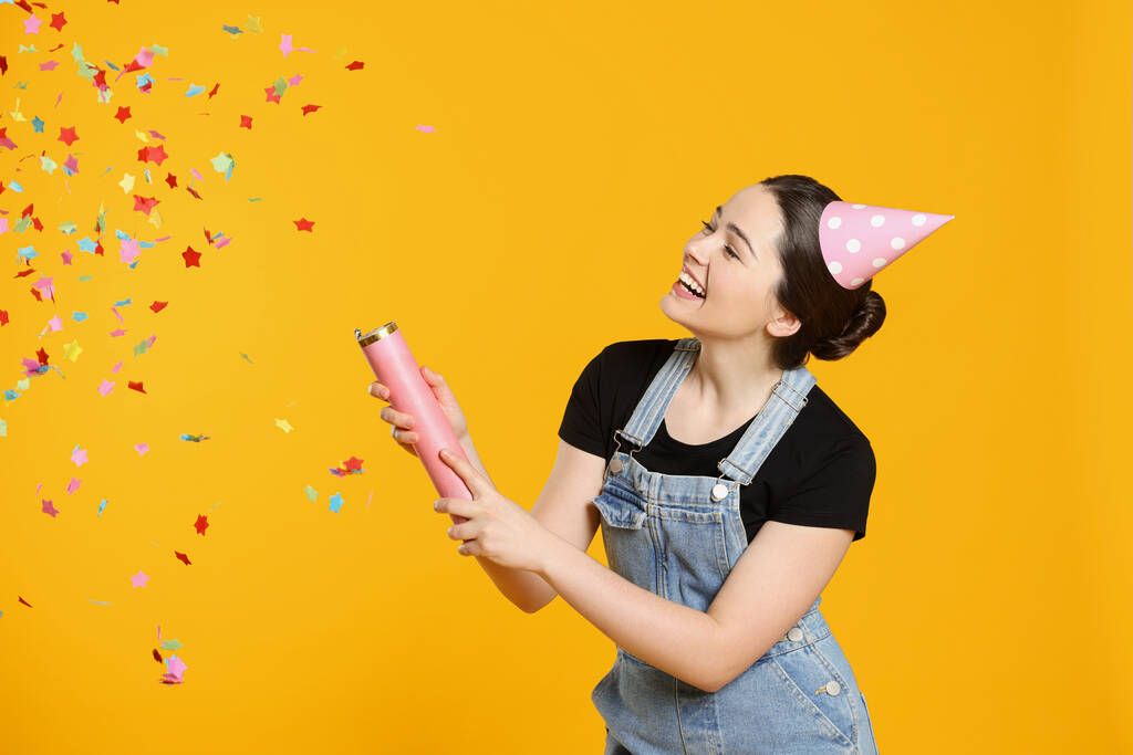 Young woman blowing up party popper on yellow background - Photo, Image
