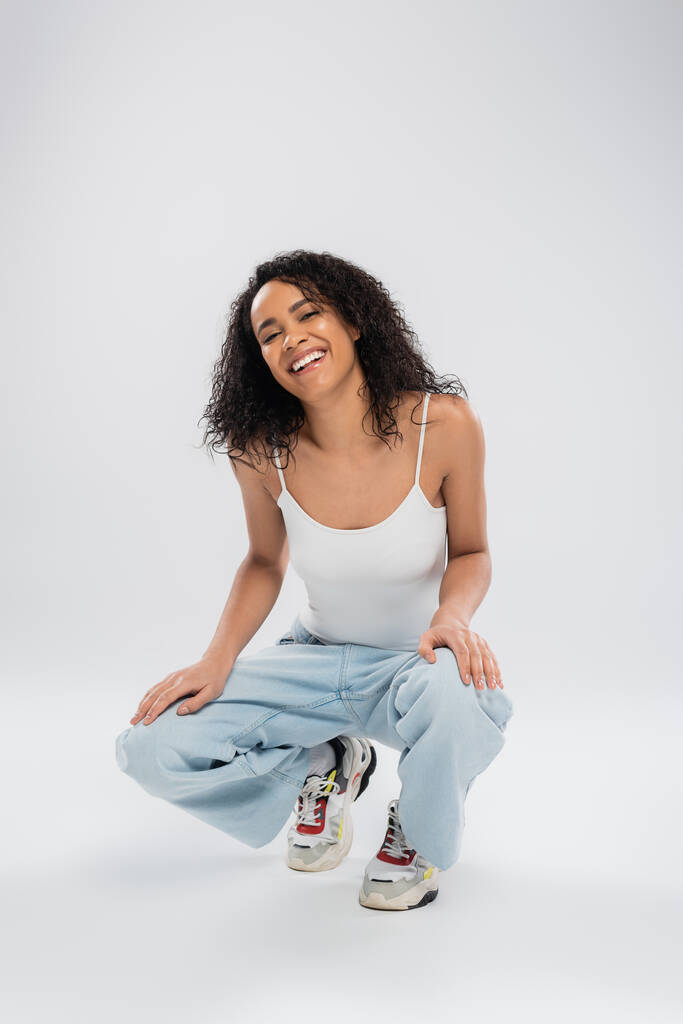 full length of overjoyed african american woman in jeans and sneakers sitting on haunches on grey background - Photo, Image