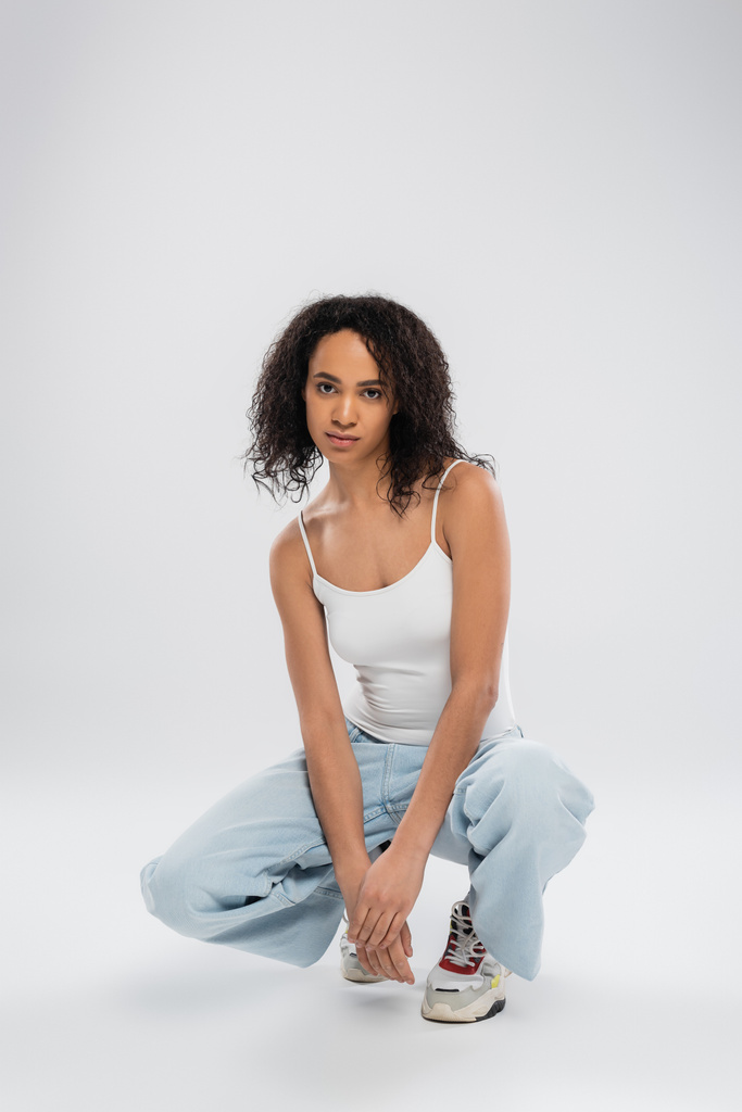 full length of african american woman in white tank top and jeans with sneakers sitting on haunches on grey background - Photo, Image