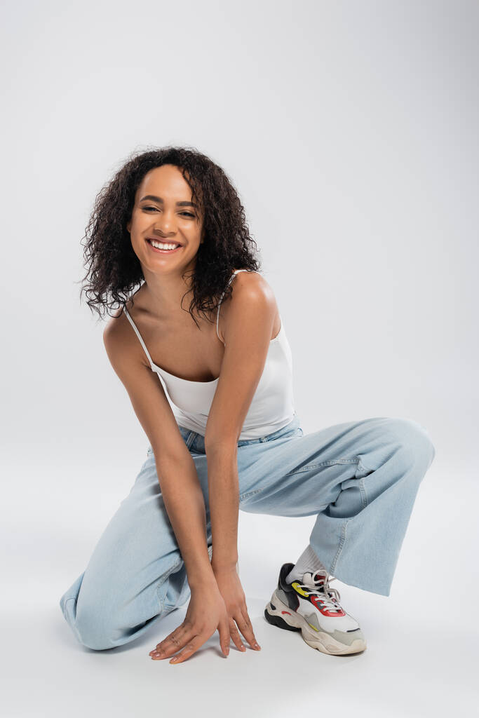 full length of excited african american woman in tank top and blue jeans posing on grey background - Photo, Image