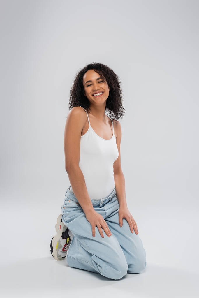 full length of pretty african american woman in jeans and white tank top sitting on haunches on grey background - Photo, Image