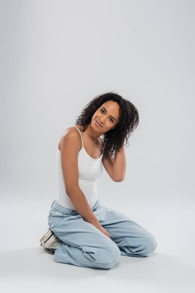 full length of positive african american woman in tank top and jeans touching hair while sitting on grey background - Photo, Image
