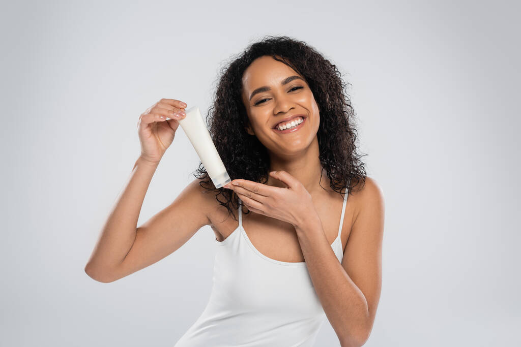 cheerful african american woman in white tank top posing with tube of cosmetic cream isolated on grey - Photo, Image