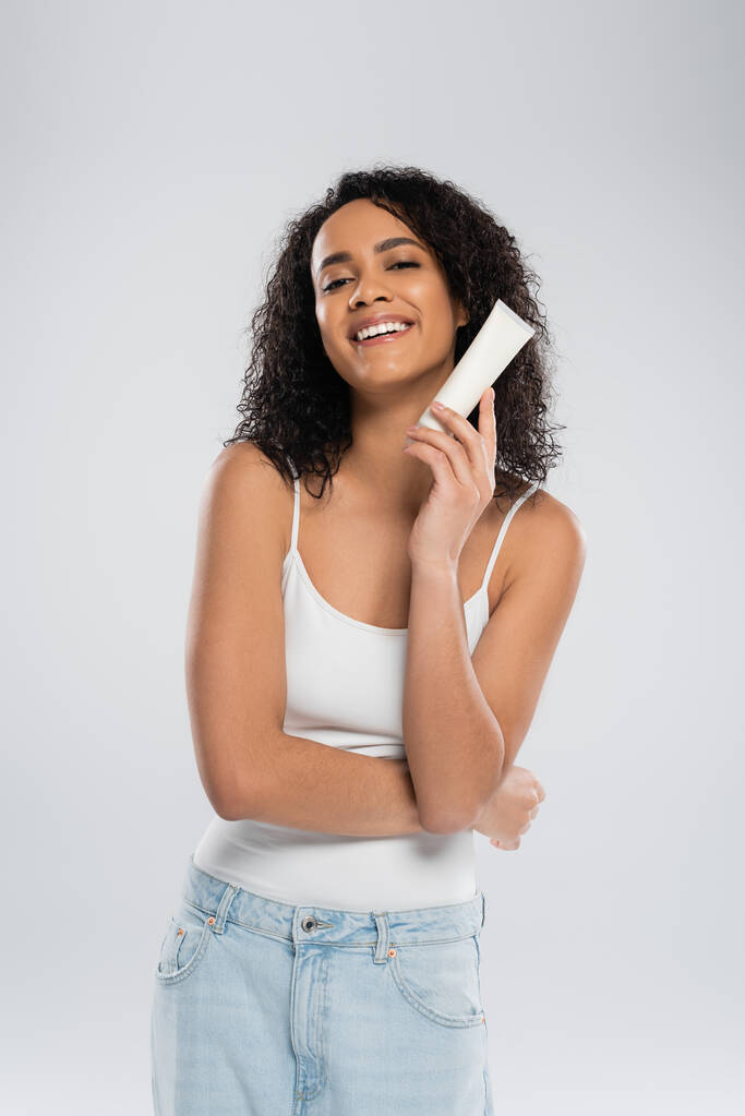 довольная африканская американка в белом топе, позирующая с тюбиком косметического крема, изолированного на сером - Фото, изображение