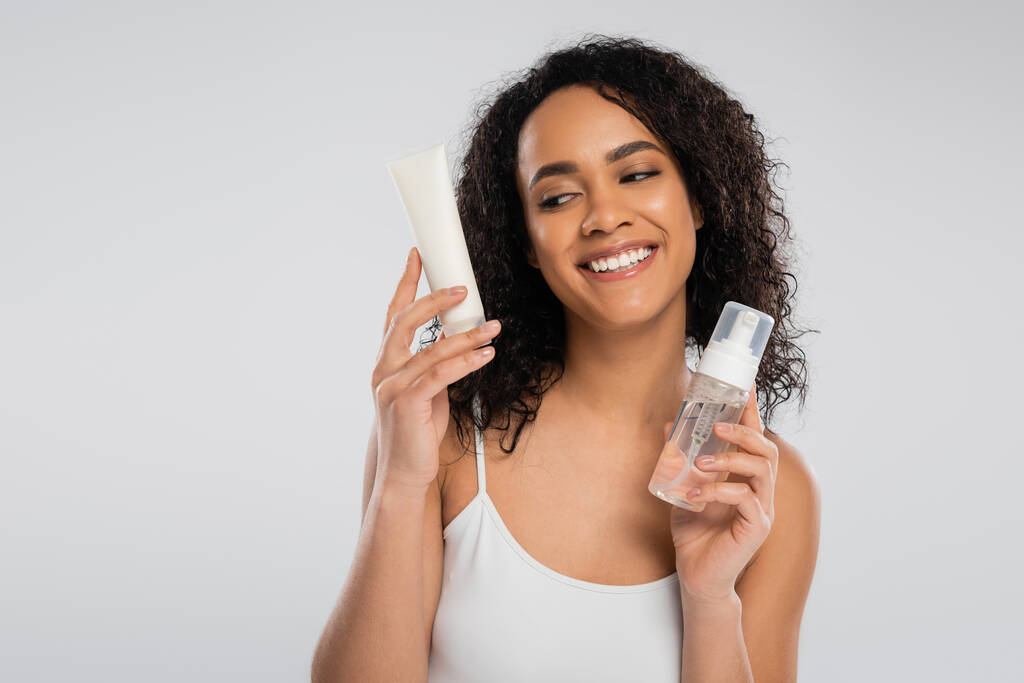 mujer afroamericana complacida sosteniendo crema cosmética y espuma facial aislada en gris - Foto, imagen