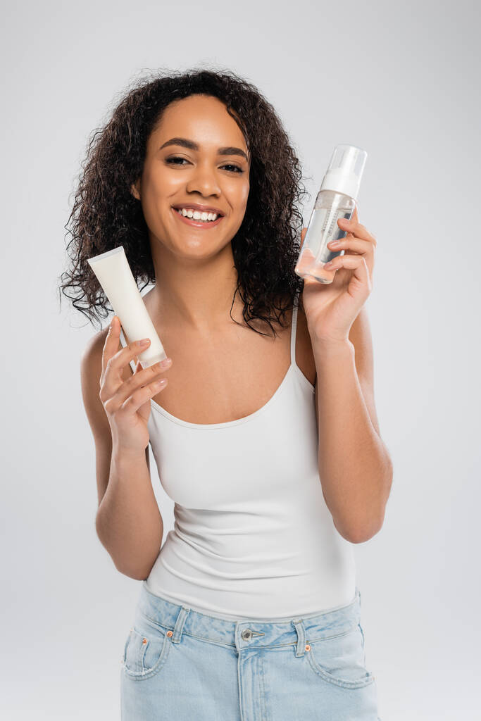 barna afro-amerikai nő fehér tartály tetején gazdaság kozmetikai krém és arc hab elszigetelt szürke - Fotó, kép