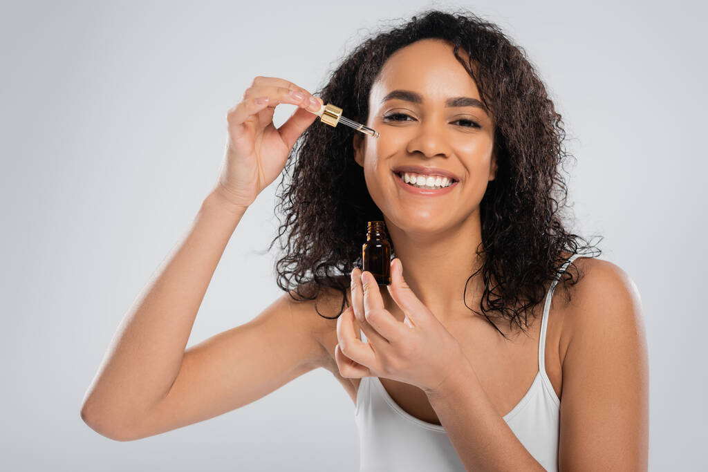 alegre mujer afroamericana aplicando suero cosmético en la cara perfecta aislado en gris - Foto, imagen