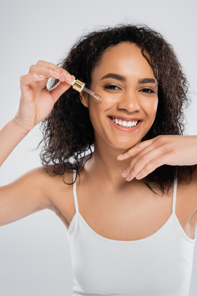zadowolona afrikańska amerykanka patrząca w kamerę i stosująca serum kosmetyczne wyizolowane na szaro - Zdjęcie, obraz