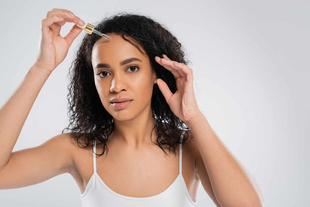 mujer americana bastante africana con piel perfecta aplicando suero cosmético aislado en gris - Foto, imagen