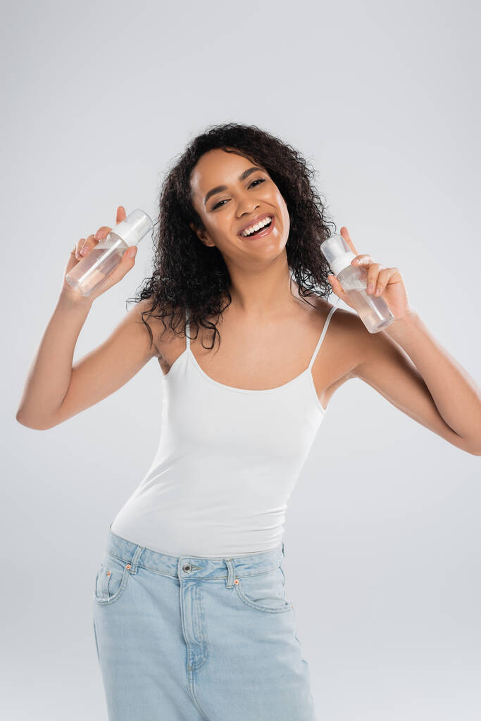 mujer afroamericana llena de alegría en camiseta blanca posando con botellas de espuma facial aisladas en gris - Foto, imagen