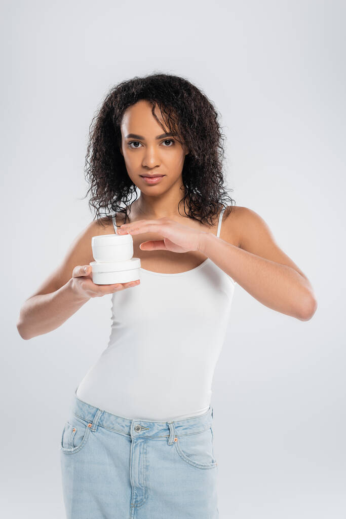 白いタンクの上の若いアフリカ系アメリカ人女性がグレーに隔離された化粧品クリームの容器でポーズをとって - 写真・画像