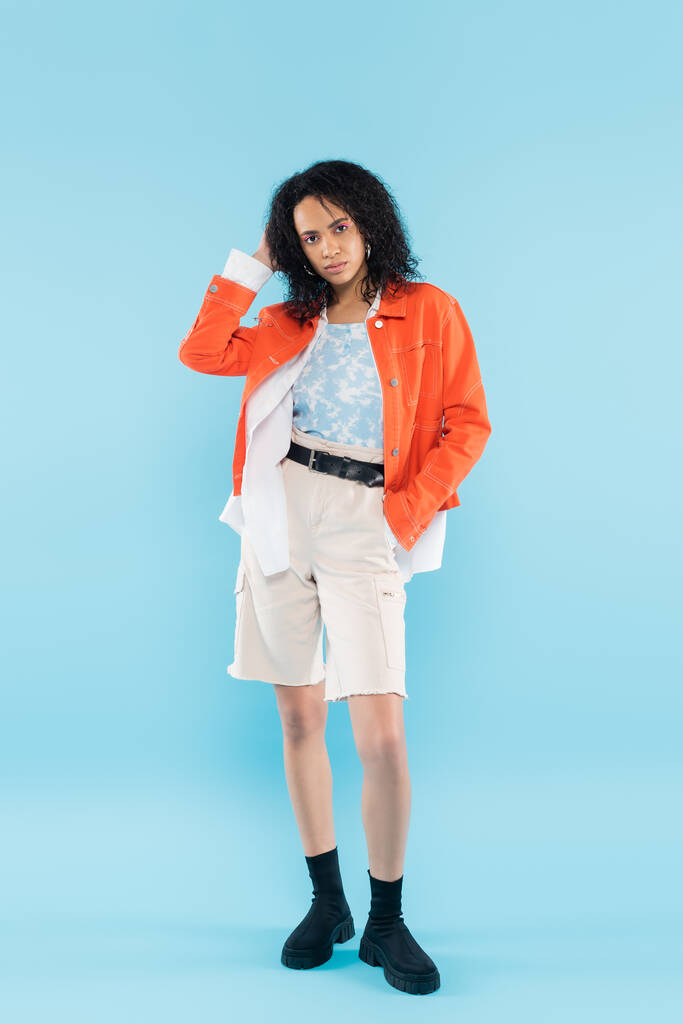 full length of stylish african american woman standing with hand in pocket of cotton shorts and touching wavy hair on blue background - Photo, Image