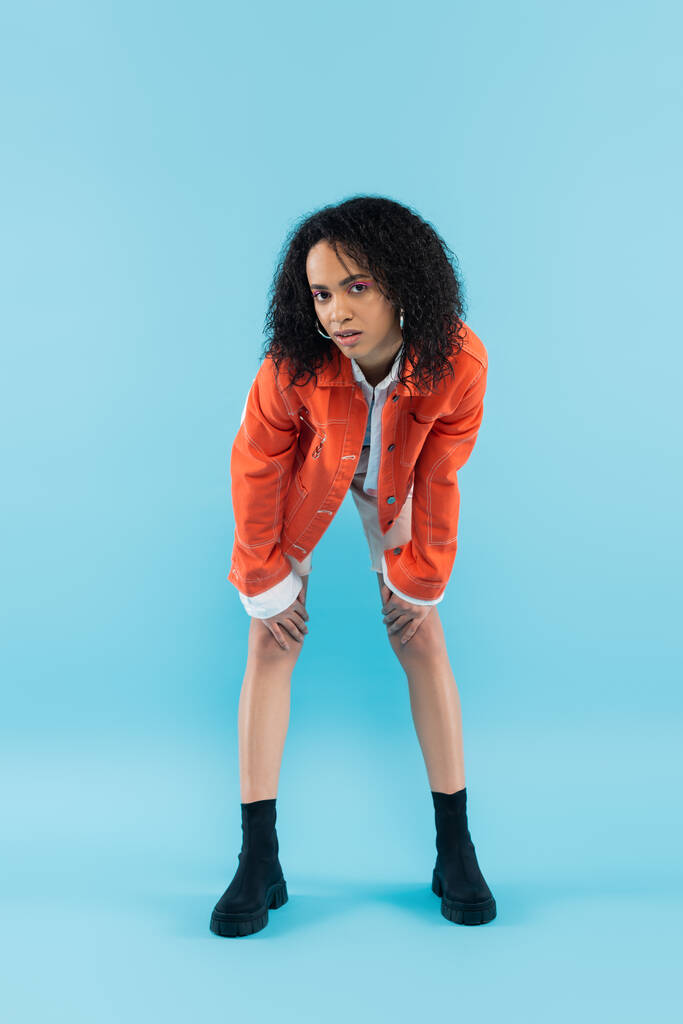 full length of curly african american woman in fashionable orange jacket posing and looking at camera on blue background - Photo, Image