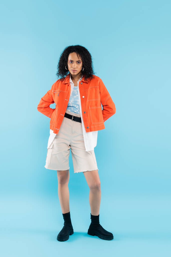 full length of trendy african american woman in white shorts and orange jacket posing with hands on hips on blue background - Photo, Image